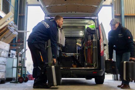 Zwei Kundendiensttechniker tragen Service-Koffer in den Firmenwagen, der in einer Halle steht.